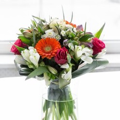 This impressively arranged bouquet, known to catch admiring glances from passersby in West Ham, is a harmonious dance of joyful colour and lush texture. At its heart rest vivacious gerbera daisies, two in a radiant orange hue and two in velvety peach, their softly arching petals embracing a distinctive chocolate-brown center. Alongside, sumptuous magenta-rose buds are artfully nestled, their swirling petals lending the arrangement an enveloping sense of romance-like a sweet gesture exchanged along the bustling Green Street. Frosty white alstroemeria, accented by clouds of petite filler flowers, scatter soft highlights and add a delicate rhythm, complemented by faint pink blooms that lend depth and nuance to the design. Thick, glossy green leaves provide a nurturing border, with broad, paddle-shaped foliage elegantly wrapped around the base, anchoring the composition in tranquil abundance. The arrangement sits within a tall, elegant glass vase, where the stems are crisply visible through the water, infusing each glance with freshness and clarity. Bathed in diffused white light reminiscent of a West Ham morning, this bouquet's radiant colours and rich textures tell a story of community warmth, professional artistry, and days filled with celebration and connection. Ideal for marking milestones, sending love, or adding a touch of contemporary beauty to a Upton Park dining room, this design is a vibrant reflection of local spirit and refined floral care.